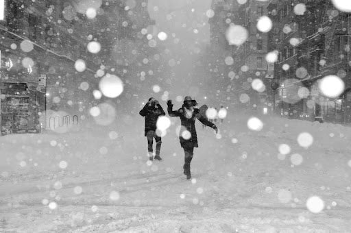 A black and white photo of two people running through a snow-covered street during a heavy snowfall, with large snowflakes falling around them.