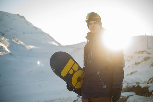 Man stands in front of the winter sunset, holdeing a snow board. | Heat Holders® Base Layers