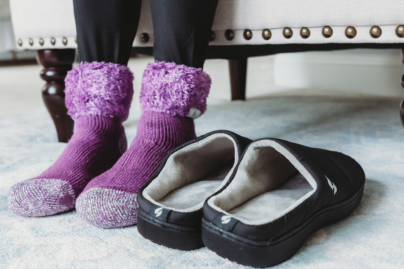  A pair of feet wearing fluffy purple ankle socks near a pair of black slippers on a carpeted floor. | Heat Holders® The Warmest Thermal Sock®