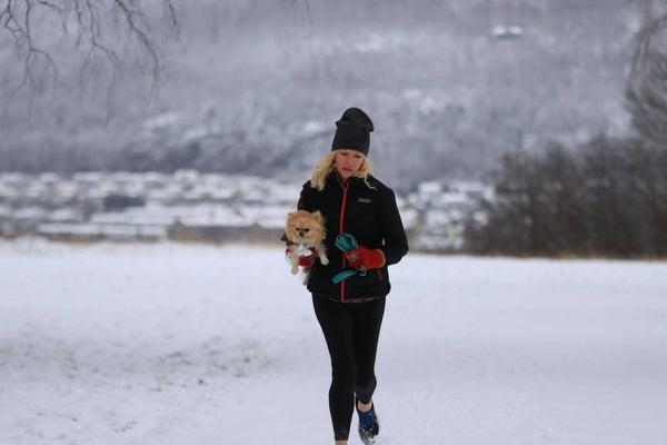 Woman in winter activewear jogging through a snowy field while carrying a small dog in her arms. Heat Holders® thermal hats.