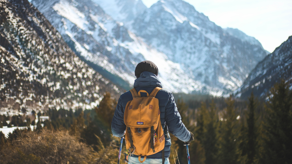 Man with a yellow backpack and trekking poles facing snow-covered mountains