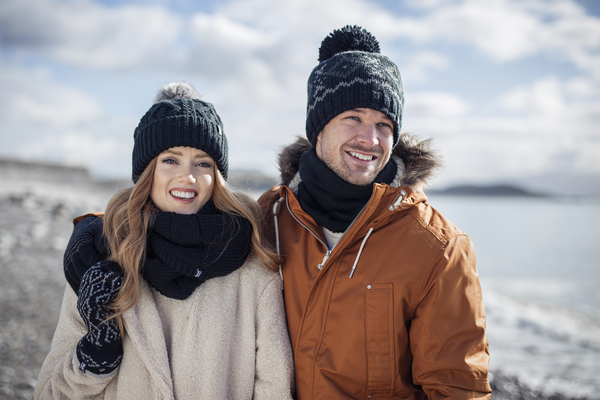 A smiling couple dressed in warm winter clothes, standing on a snowy beach. The woman wears a beige coat, black knit hat, and gloves, and the man is in an orange jacket with a fur-lined hood, neck warmer, and a matching knit hat