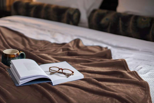 Reading glasses on top of a book beside a coffee mug, all on a luxurious Heat Holders® throw on a bed.