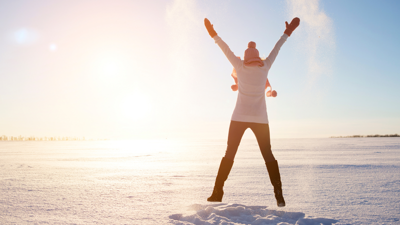 Woman in winter clothing outside in the snow jumping with arms up.